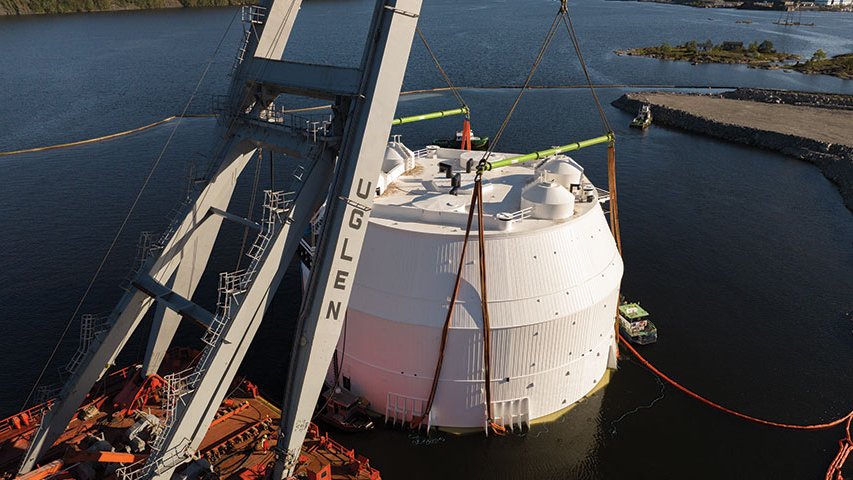 Großer zylindrischer Behälter wird mit Kran ins Wasser gehoben, befestigt mit Gurten, Arbeitsplattform daneben