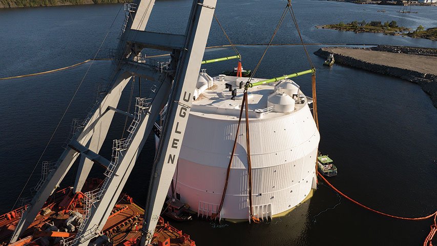 Large cylindrical tank lifted into water by crane, secured with straps, work platform alongside