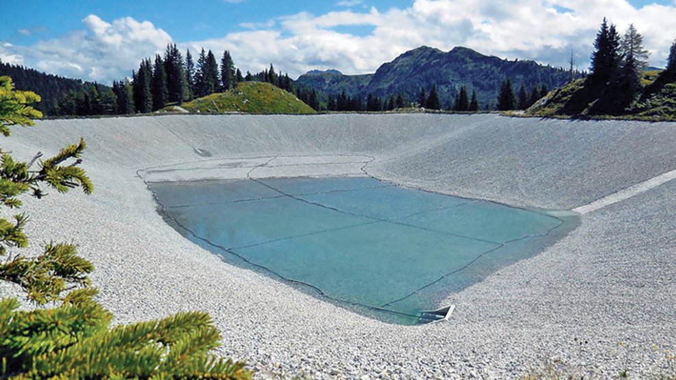Man-made water basin with a bright liner, surrounded by mountain scenery, woodland areas, and gravel embankments.