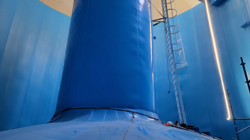 Innenansicht eines großen blau ausgekleideten Behälters mit zentraler Säule, Leiter und heller Beleuchtung.