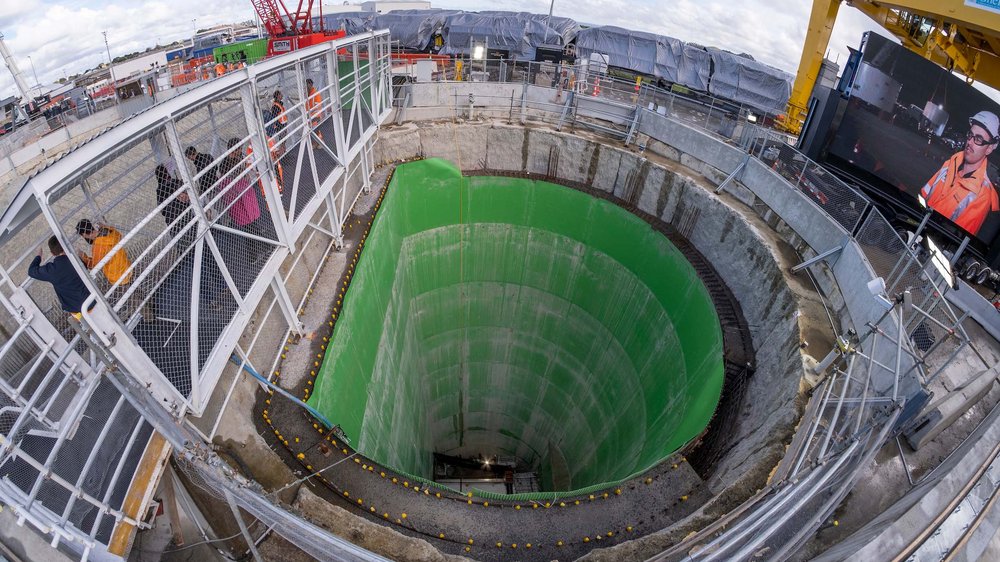 Industriebaustelle mit tiefem Schacht, Metallgeländern, Arbeitsplattformen und sichtbaren Anlagen im Hintergrund.