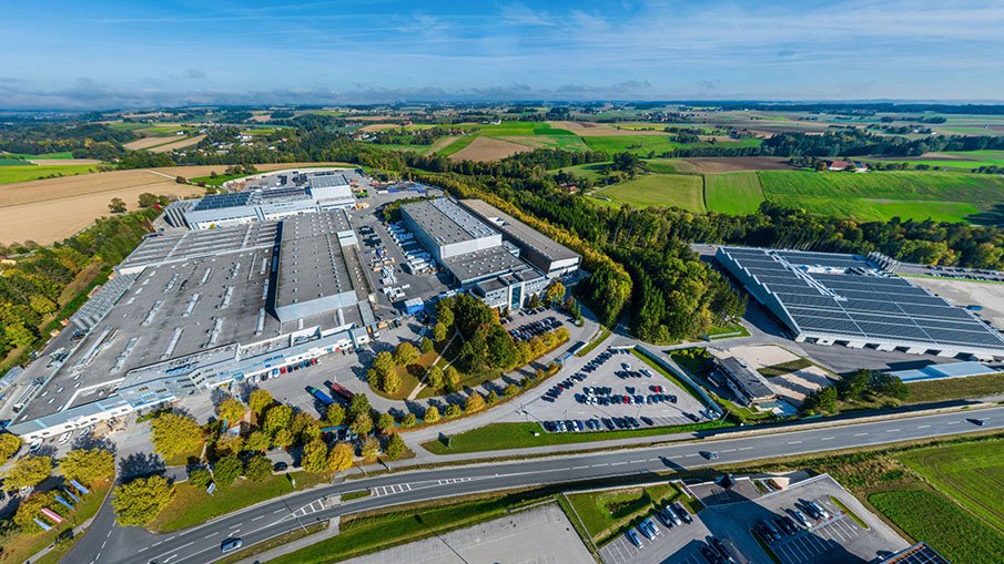 Luftaufnahme eines Produktionsstandorts mit mehreren Industriehallen, Solaranlagen und Parkflächen in ländlicher Umgebung