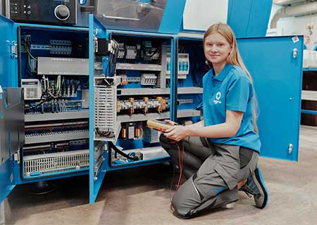 Apprentice working on manufacturing equipment with open access doors, technical parts and tools visible