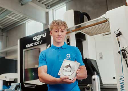 Apprentice holding finished workpiece in front of manufacturing equipment and machine workstation