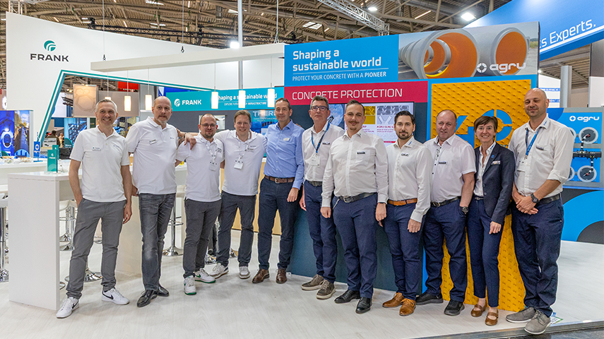 Mitarbeitende präsentieren sich gemeinsam vor Messestand mit technischen Visualisierungen und Beschriftungen