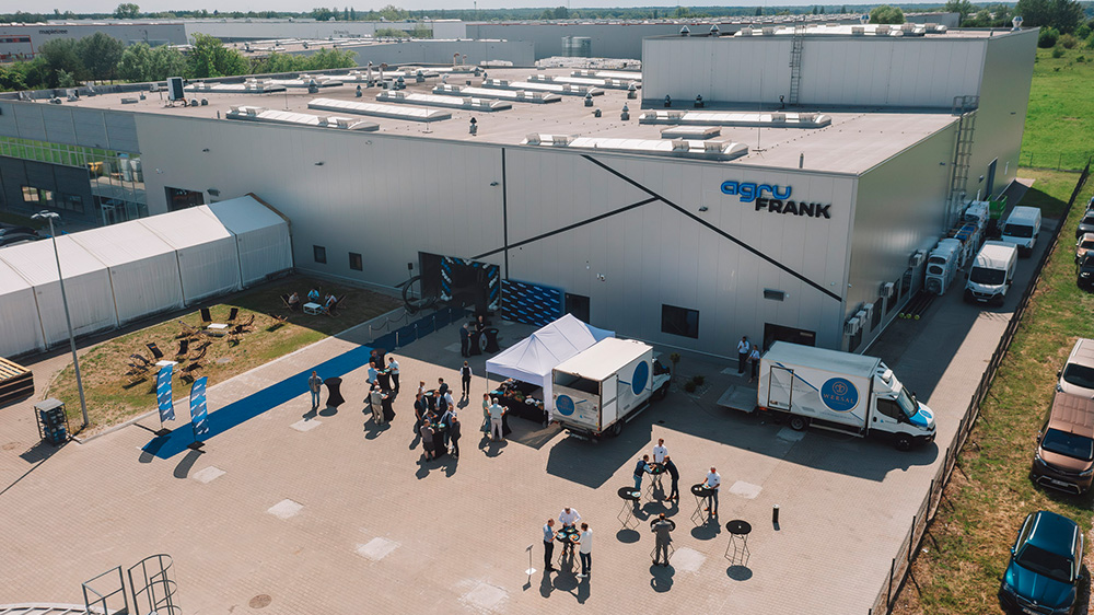 Large warehouse building with “AGRU FRANK” sign, event tents, delivery trucks and small groups of visitors outside