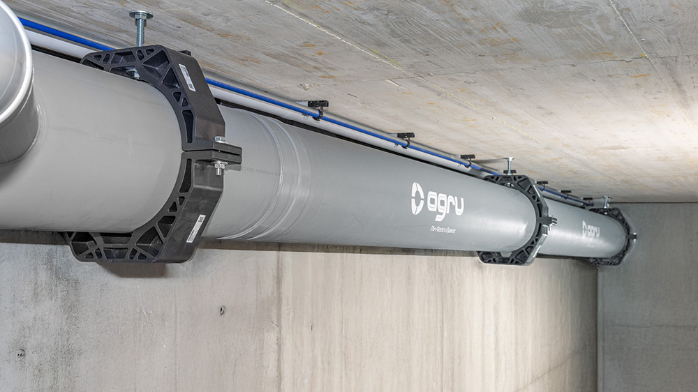 Gray plastic pipe with AGRU logo, secured to a concrete ceiling using black pipe clamps and threaded rods.
