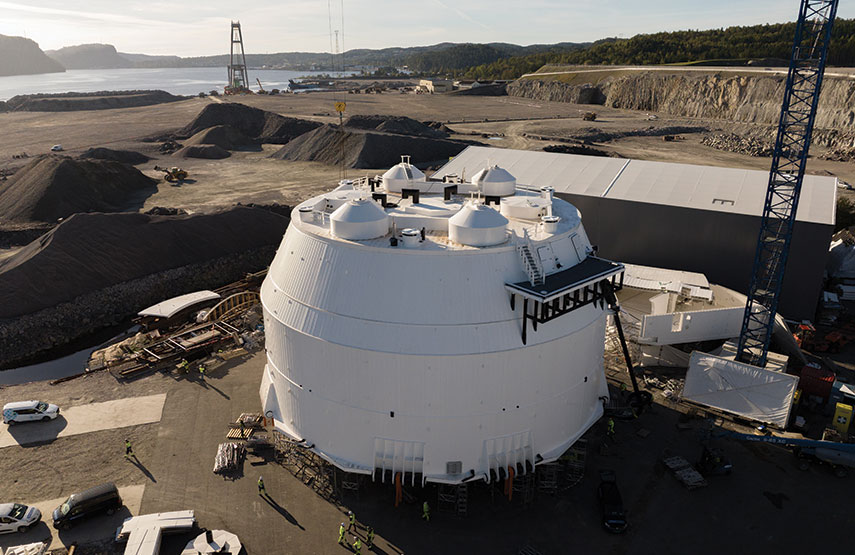 Large white circular tank installed on construction site, crane nearby, vehicles and workers visible around