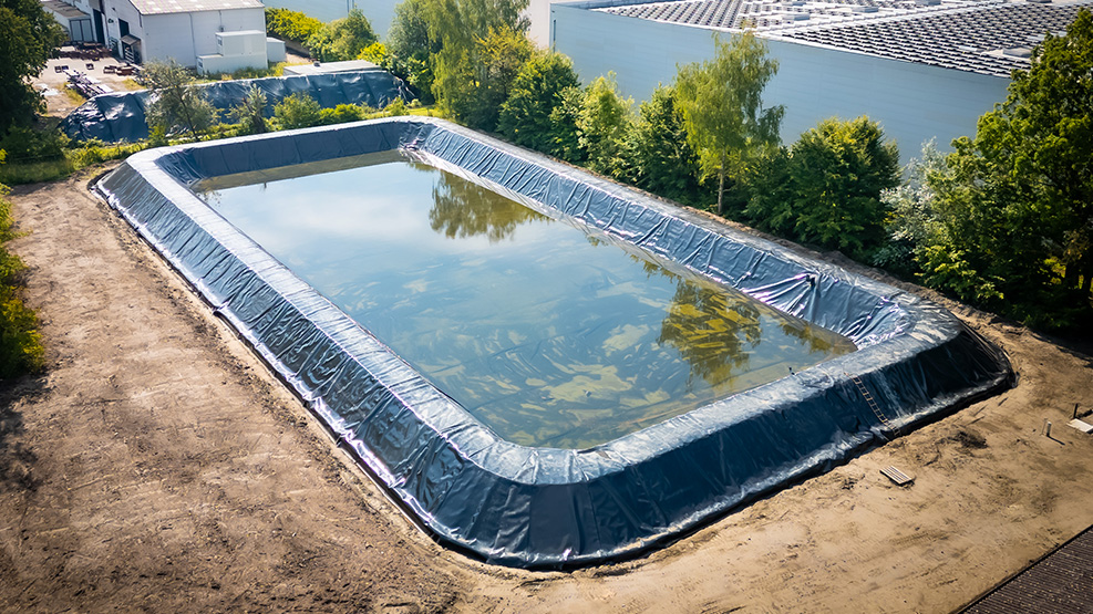 Rechteckiges Wasserbecken im Freien, ausgekleidet mit dunkler Folie, umgeben von Erde und Grünflächen neben Gebäuden.