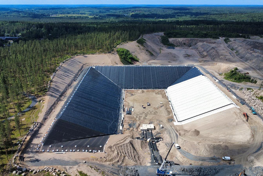 Großes Speicherbecken im Bau mit schwarzer und weißer Folienauskleidung, umgeben von Wald und Erdarbeiten.