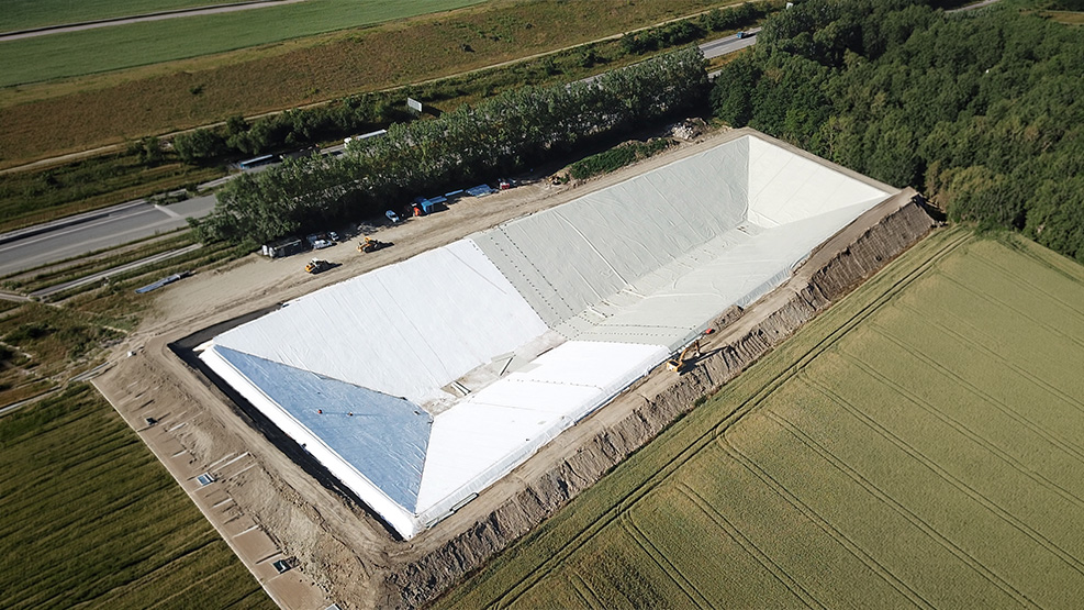Luftaufnahme eines großen Speicherbeckens mit weißer Folienauskleidung, umgeben von Feldern, Wald und Zufahrtswegen.