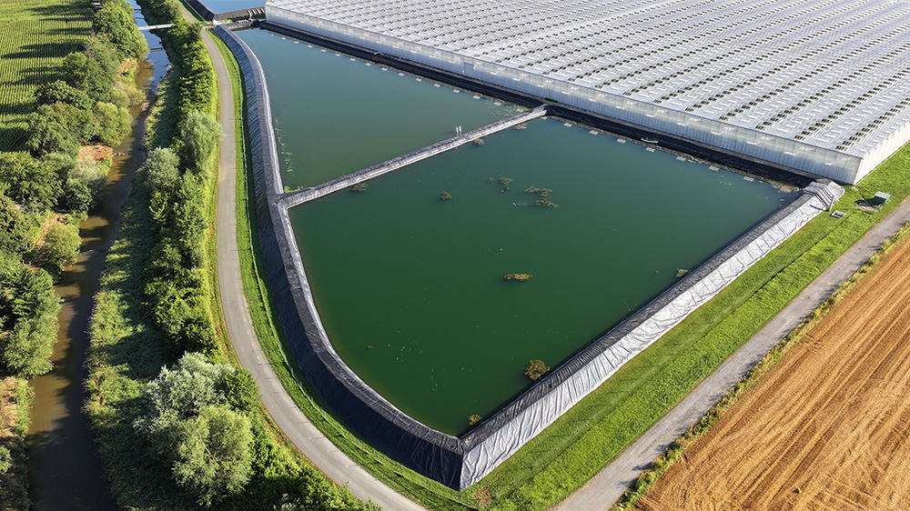 Luftaufnahme eines mit Wasser gefüllten Speicherbeckens mit dunkler Abdichtung entlang einer Gewächshausanlage.