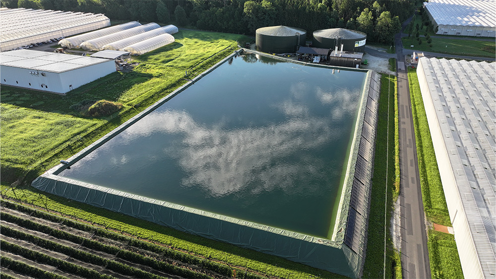 Folienabgedichtetes Wasserbecken in einer Gartenbauanlage, umgeben von Hallen, Wegen und bewachsenen Flächen.