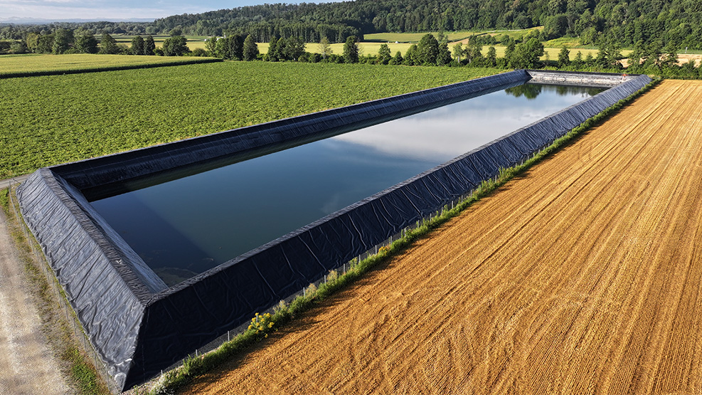 Inmitten landwirtschaftlicher Flächen liegt ein langes Speicherbecken mit dunkler Abdichtung und ruhiger Wasseroberfläche.